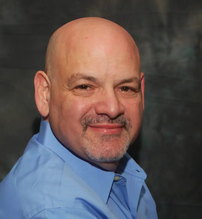 Marcel Lashover wears a blue shirt, smiling in front of a dark background.