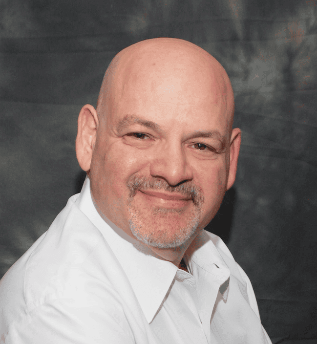 A bald man with a goatee and wearing a white shirt smiles against a dark, textured background.