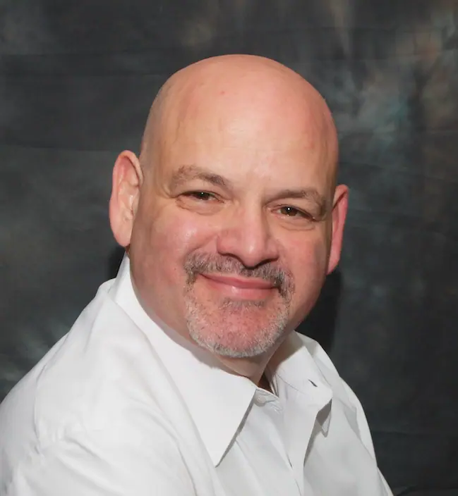 Marcel Lashover wearing a white shirt smiles against a dark, textured background.