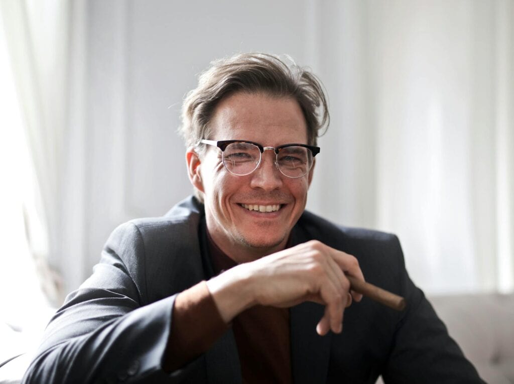 Cheerful businessman in glasses and suit holding a cigar, indoors.