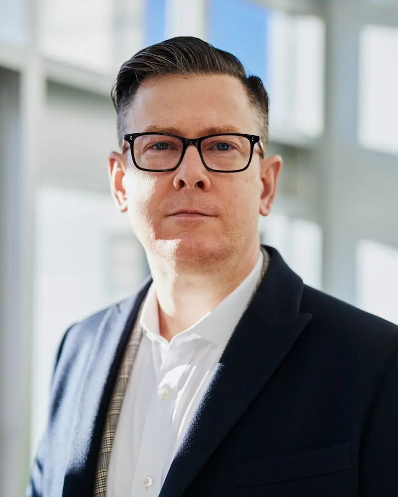 A man wearing glasses, a white shirt, and a dark blazer stands indoors in front of windows with natural light.