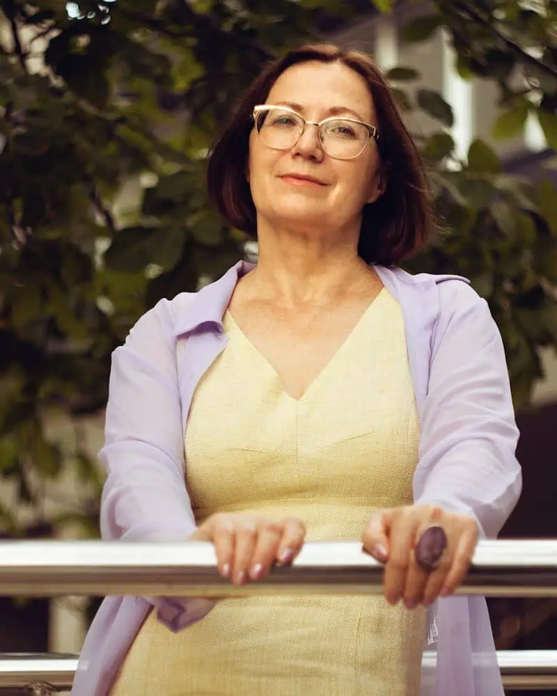 A woman with glasses and short brown hair stands outdoors, wearing a light yellow dress and a sheer lavender shawl, resting her hands on a metal railing.