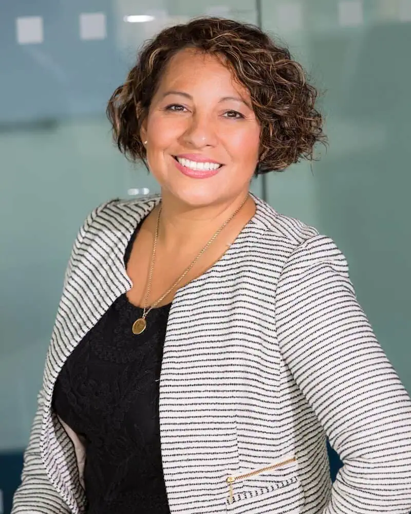 A woman with short curly hair, wearing a striped blazer over a black top, smiles while standing in an office setting.