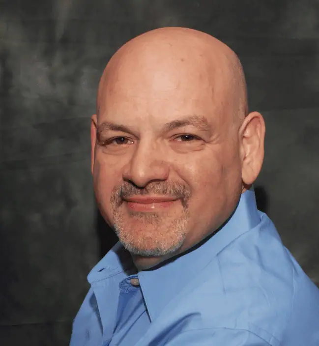 A middle-aged bald man with a goatee and mustache is wearing a light blue collared shirt and smiling slightly at the camera against a dark background.