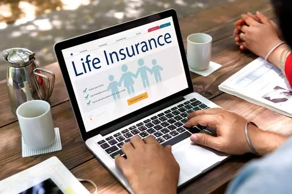 A person uses a laptop displaying a life insurance webpage with family icons, check marks, and an "Apply for a quote" button, sitting at a wooden table outdoors.