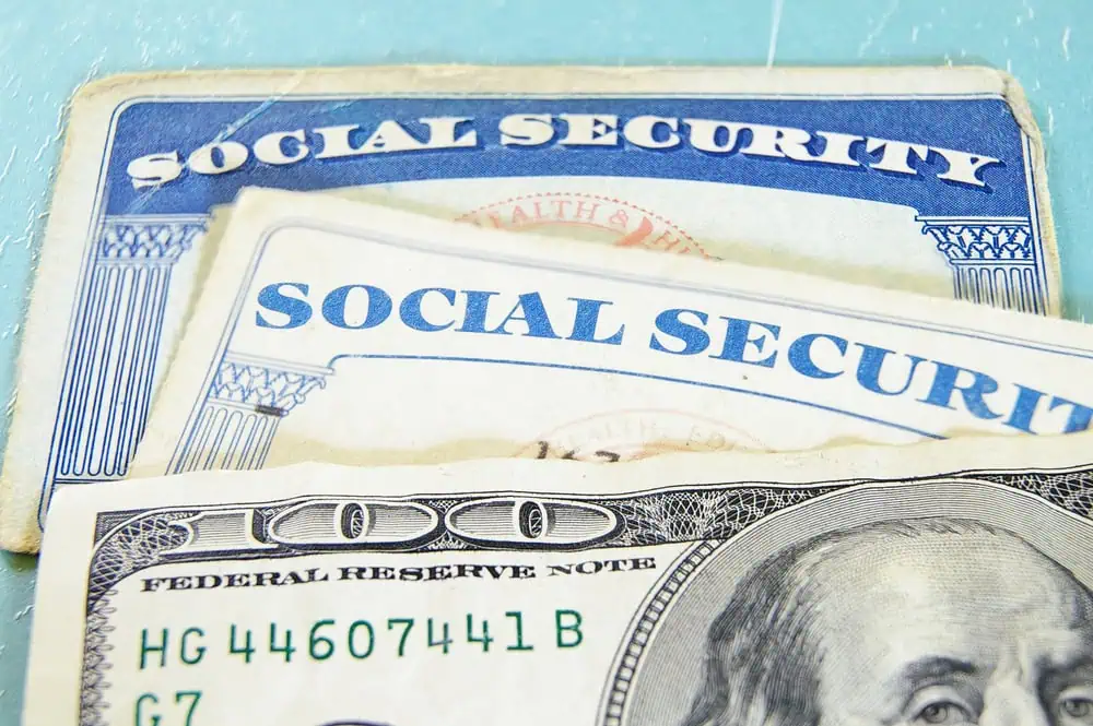 Three Social Security cards are stacked above a one hundred dollar bill on a light blue surface.