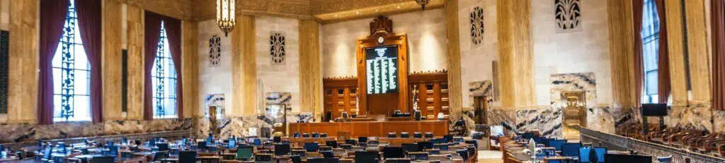 A large legislative chamber with rows of desks, high ceilings, tall windows, and marble walls—where key issues like the terminal illness tax deduction and Act 251 Louisiana are discussed beneath a central board at the front.