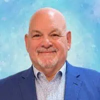 Bald man with a gray goatee wearing a blue blazer and checked shirt, smiling in front of a blue, softly textured background.